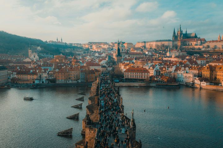 A scenic view of the Charles Bridge in Prague, Czech Republic, crowded with people and surrounded by historic buildings with orange rooftops. The Vltava River flows beneath, and Prague Castle is visible in the distance atop a hill.