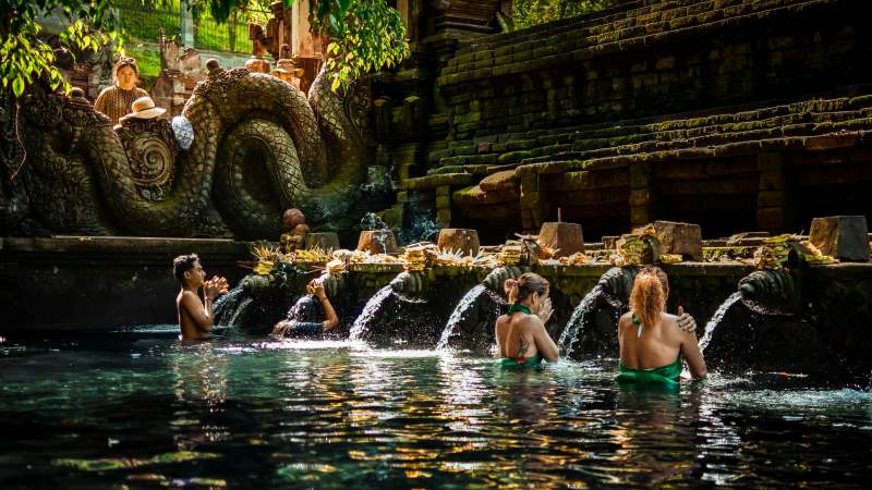 People participating in the sacred purification ritual at Tirta Empul Temple in Bali, with intricate stone carvings and gushing water spouts visible. A significant spiritual site among temples to visit in Bali.