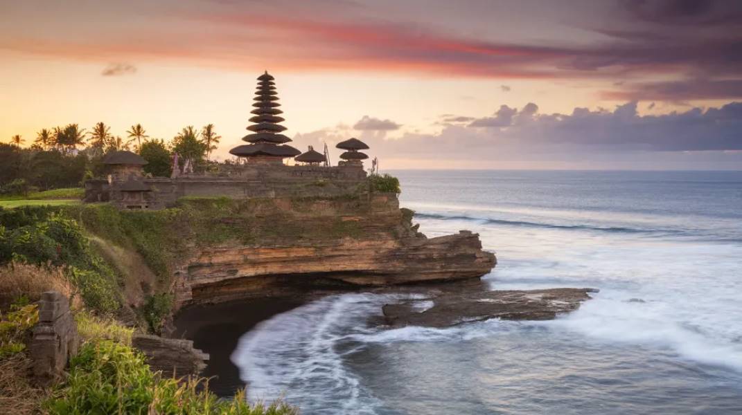 Pura Luhur Uluwatu Temple perched dramatically on a cliff overlooking the ocean at sunset, showcasing its multi-tiered meru structure against a vibrant sky, a prominent temple to visit in Bali."