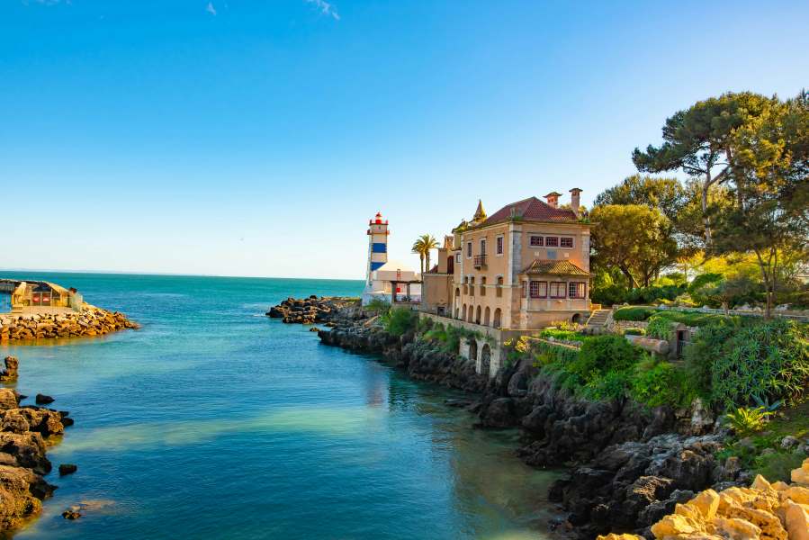 Scenic view of Cascais, Portugal with a lighthouse, historic villa, and rocky coastline by the Atlantic Ocean.