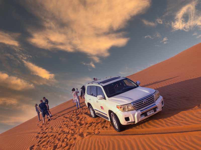 Private desert safari in Dubai with tourists exploring the Lahbab Red Dunes beside a 4x4 Land Cruiser at sunset.